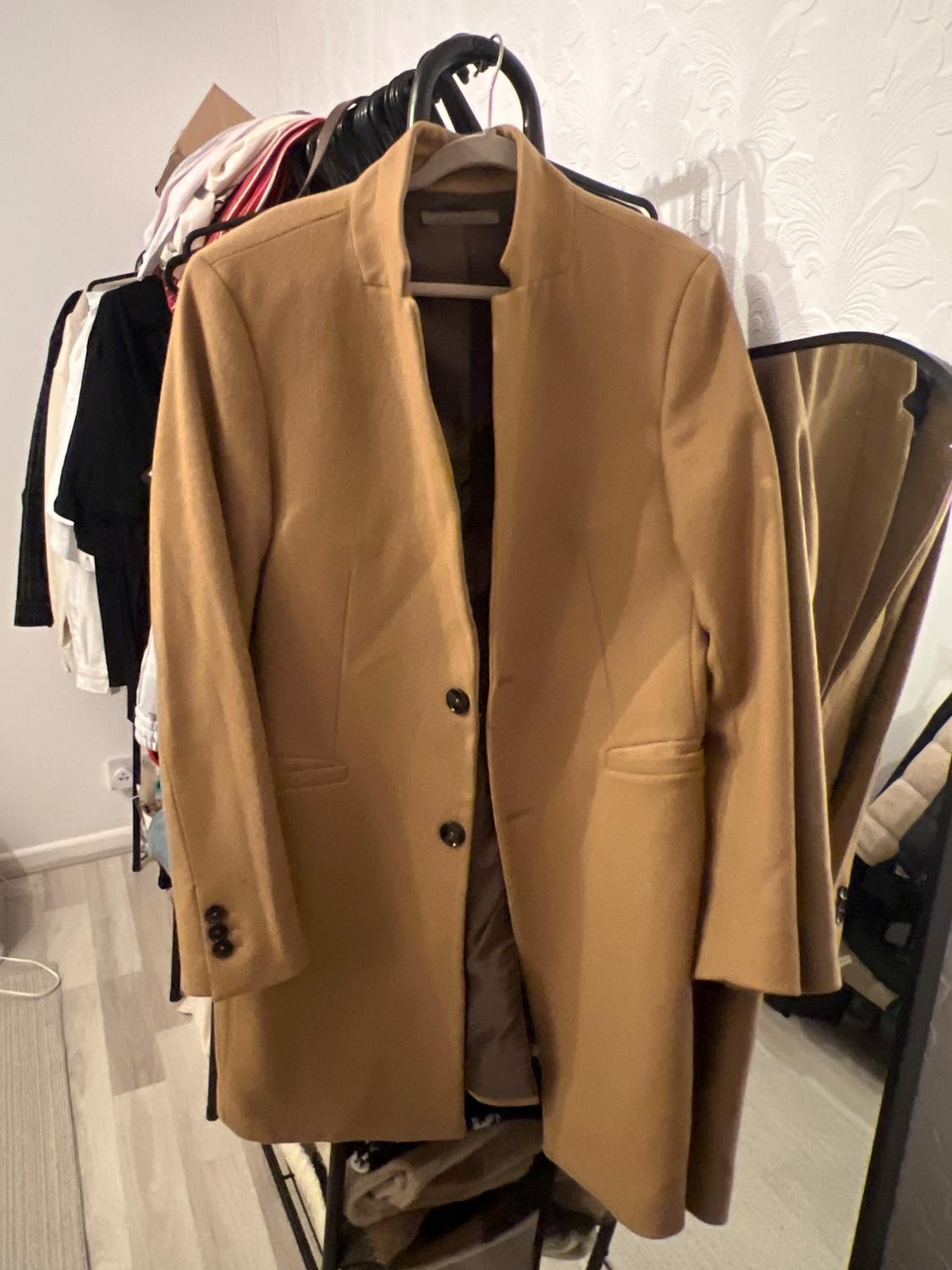 Camel coat hanging on a clothes rail with a background of various clothes and a mirror. Bedroom setting with light wooden flooring.