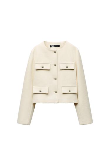 Long-sleeved white jacket with four front pockets and metal buttons. Visible label with "ZARA WOMAN" text.