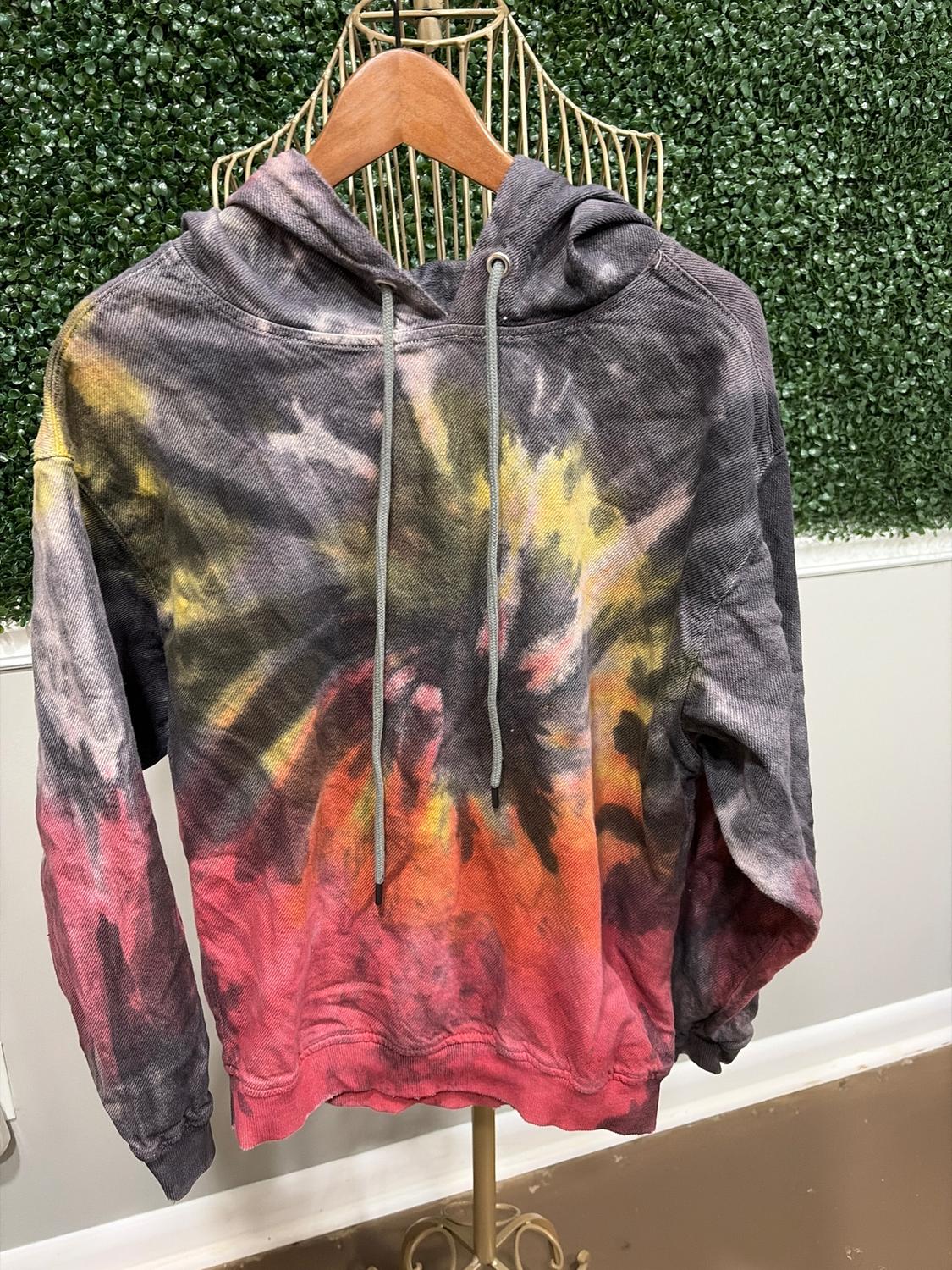 Vibrantly colored tie-dye hoodie, hanging on a wooden hanger in front of a green leafy background.