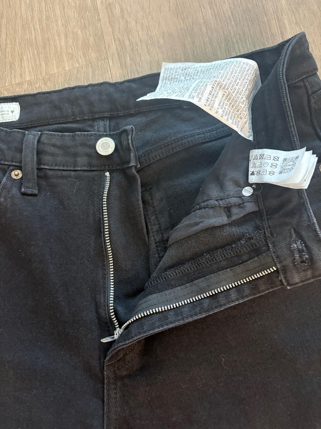 Black jeans with an open zipper and a silver button. The inner care labels are shown on a light wood background.