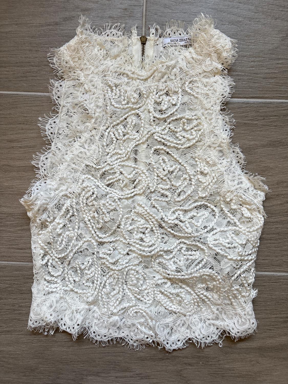 Sleeveless white lace top with embroidered details and floral texture, gold zip fastening at the back, on a wooden background.