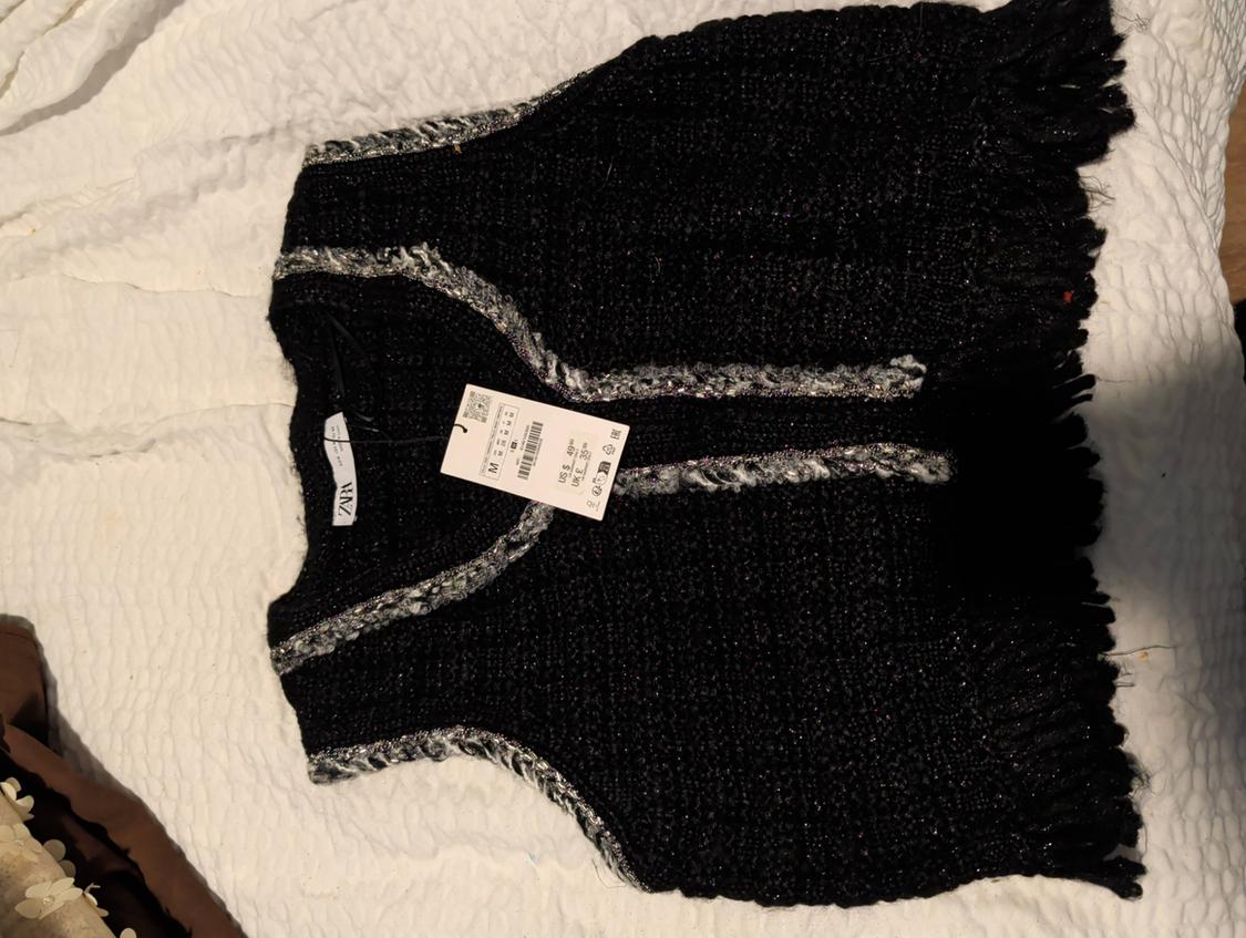 Black vest with fringes and gray details on a white bed. Visible tag with size and price information.