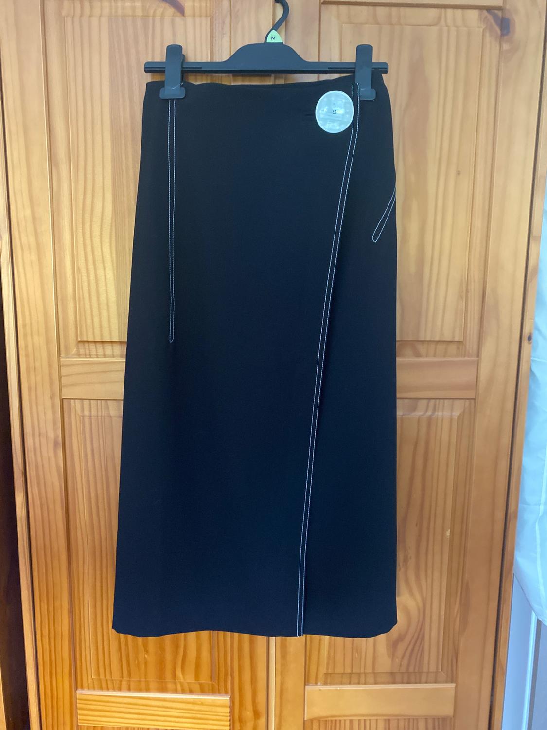 Long black skirt with visible white stitching, hanging on a black hanger over a wooden door. Round tag at the waist.