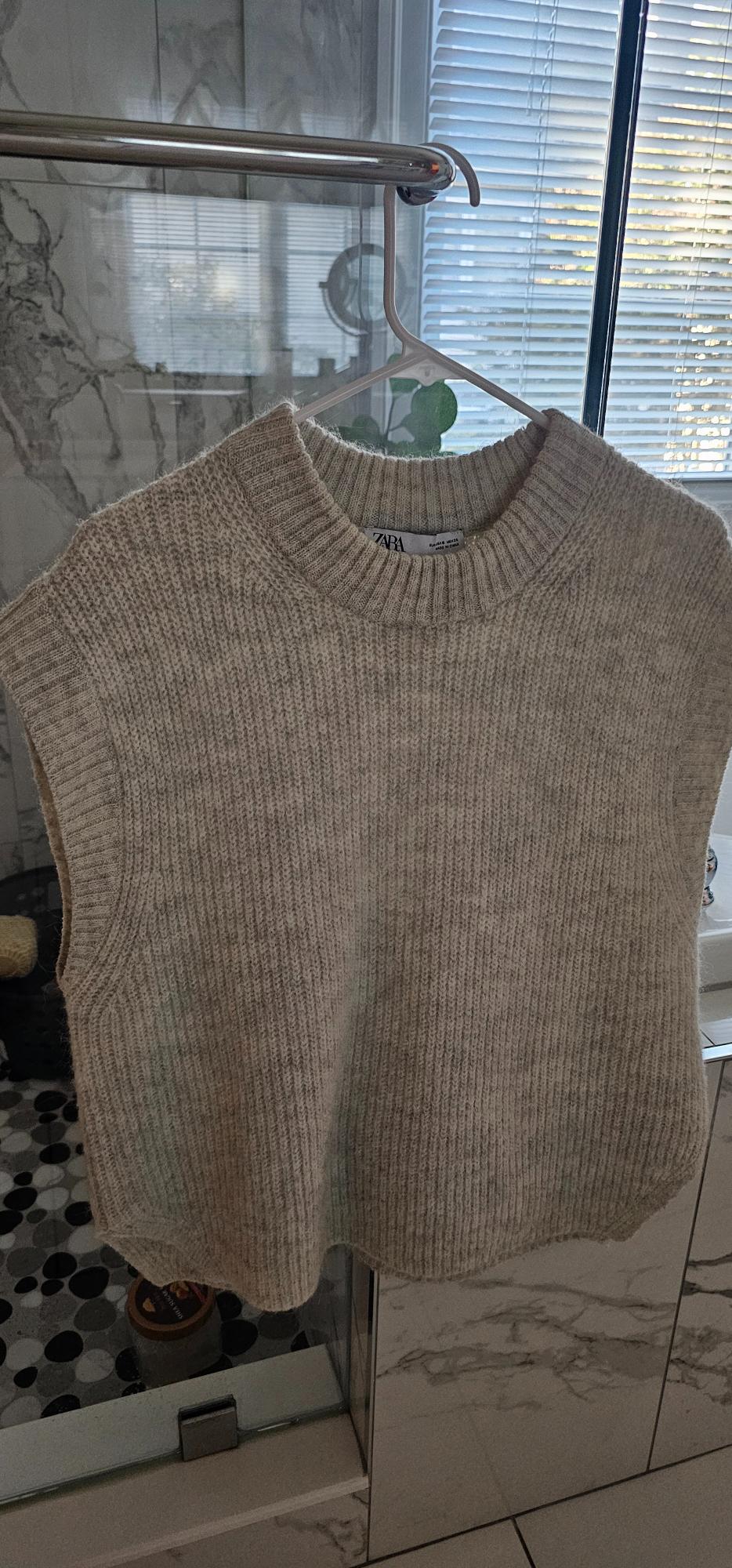 Sleeveless knit vest in heathered beige with a crew neck, hanging on a hanger in a bathroom. The Zara brand label is visible.