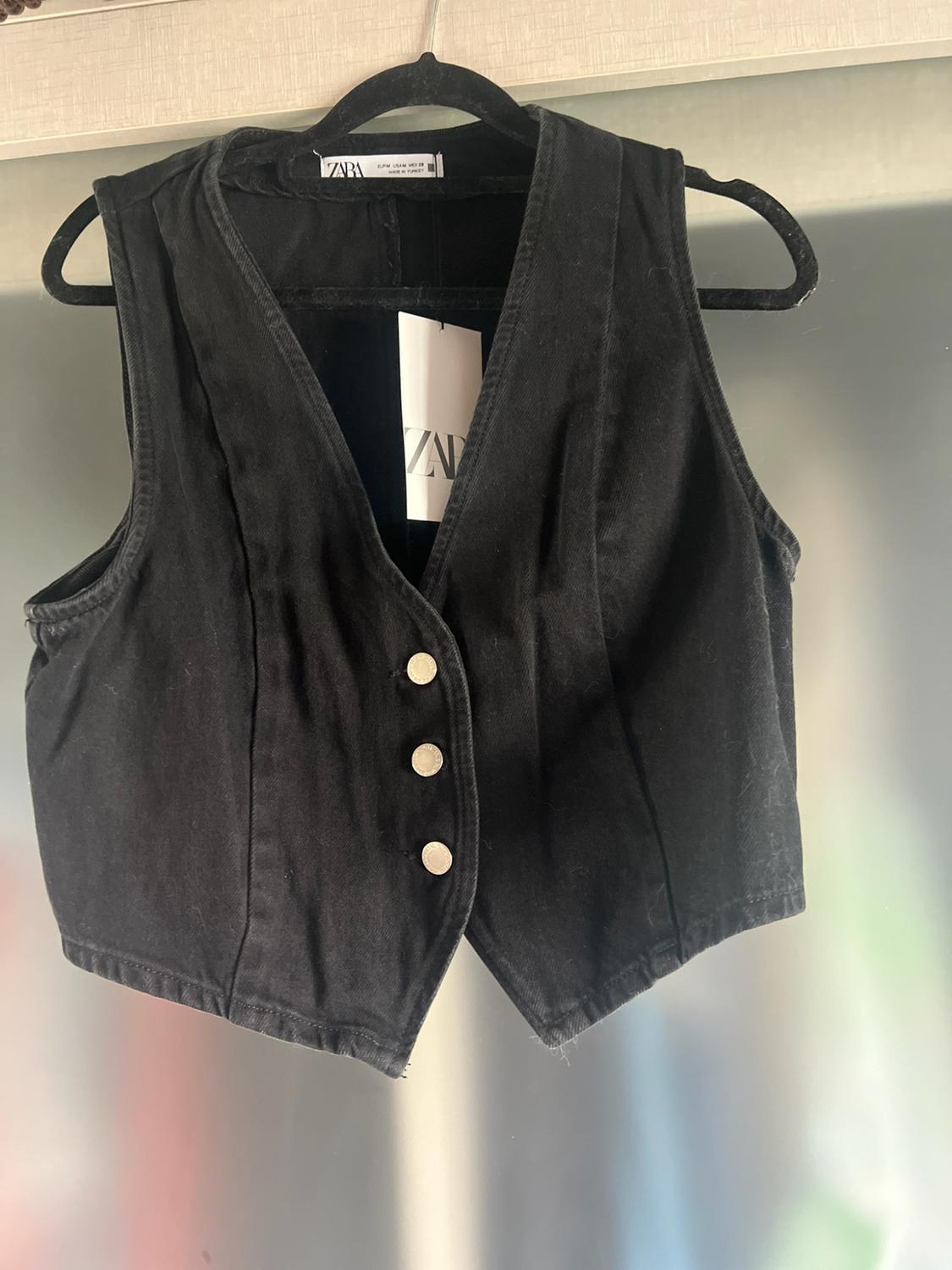 Black denim waistcoat with a V-neck and three buttons hanging on a coat hanger. Inside, a label bearing the text ZARA can be seen.