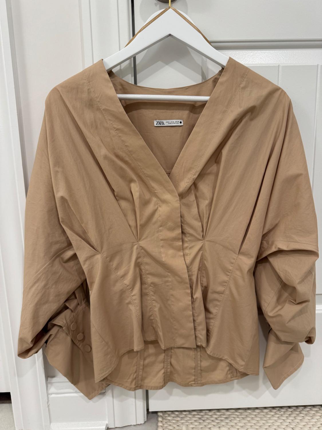 Beige long-sleeved blouse with pleats, hanging on a white hanger in front of a white door. Visible label: ZARA.