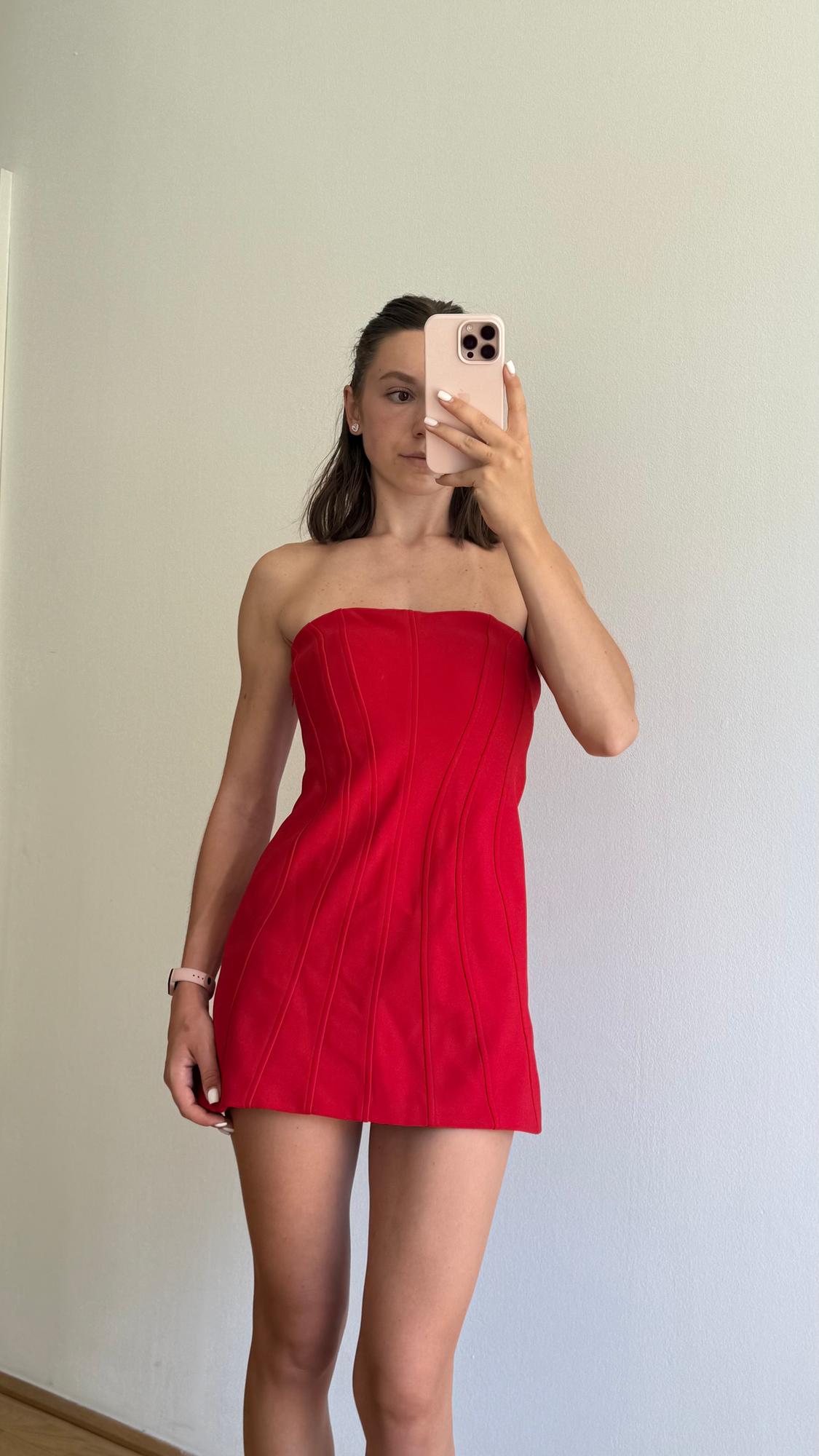 Woman in a strapless red dress, taking a selfie in front of a mirror. White background and watch on her left wrist.