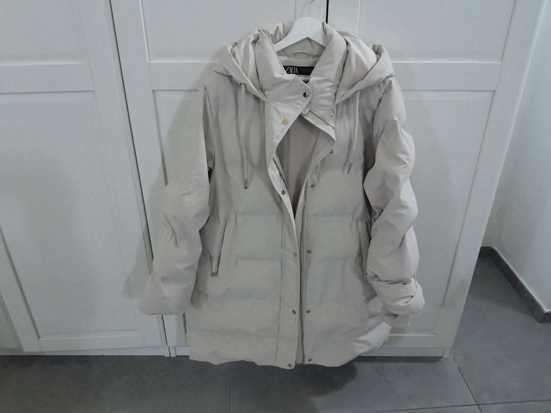 Beige padded coat with a hood hanging on a white hanger in front of a white wardrobe on a grey tiled floor.