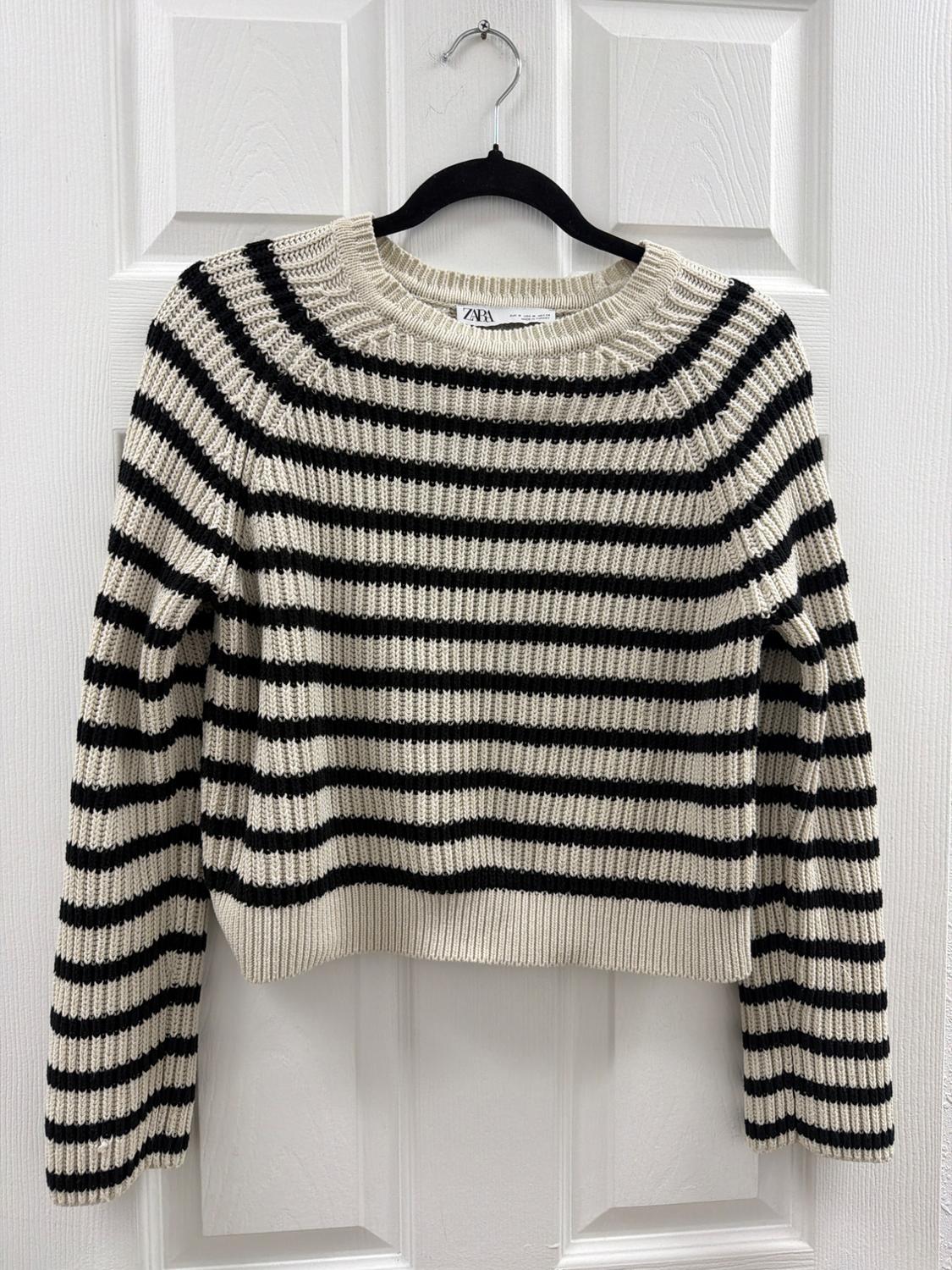 Knitted jumper with black and white horizontal stripes hanging on a hanger on a white door. Label with the text ZARA.