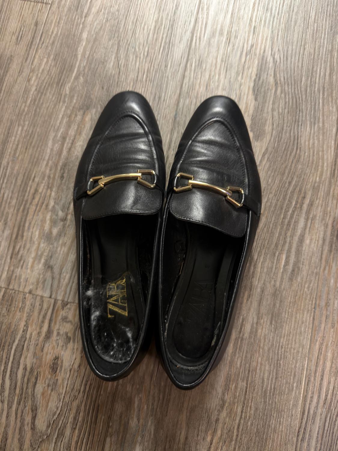 Black leather shoes with a gold detail on the top, placed on a wooden floor. Visible text: "ZARA".