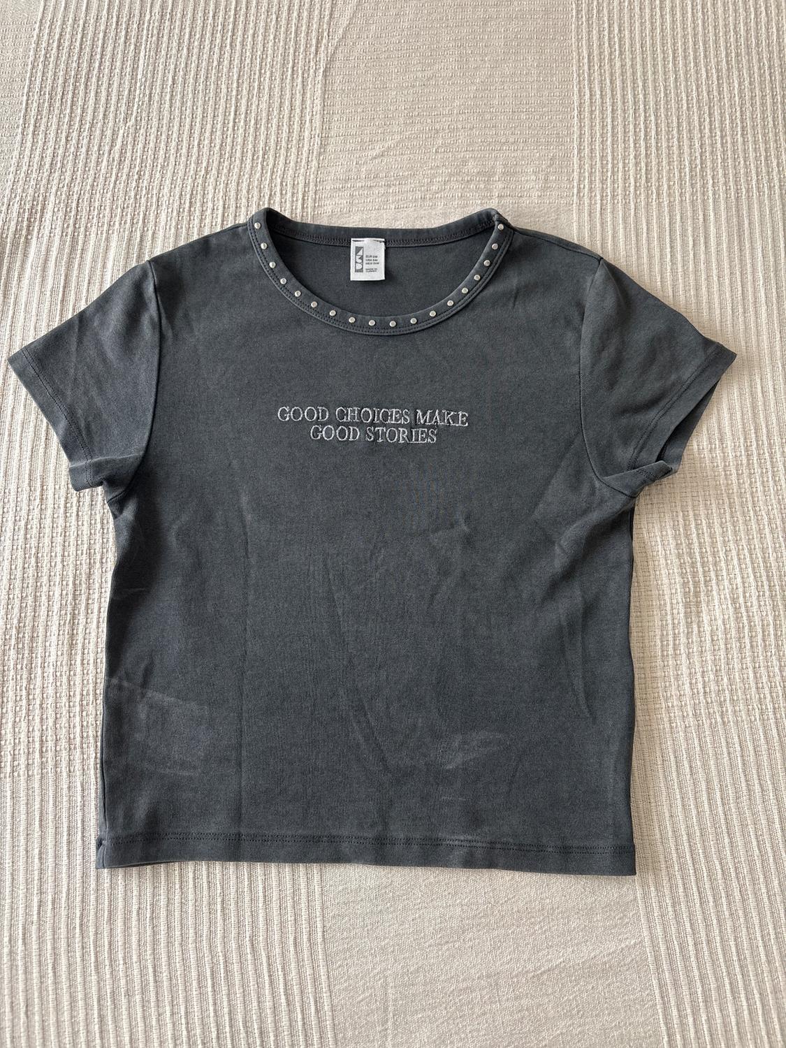 Grey T-shirt with studs on the collar and the text "GOOD CHOICES MAKE GOOD STORIES" in the centre, on a beige surface.