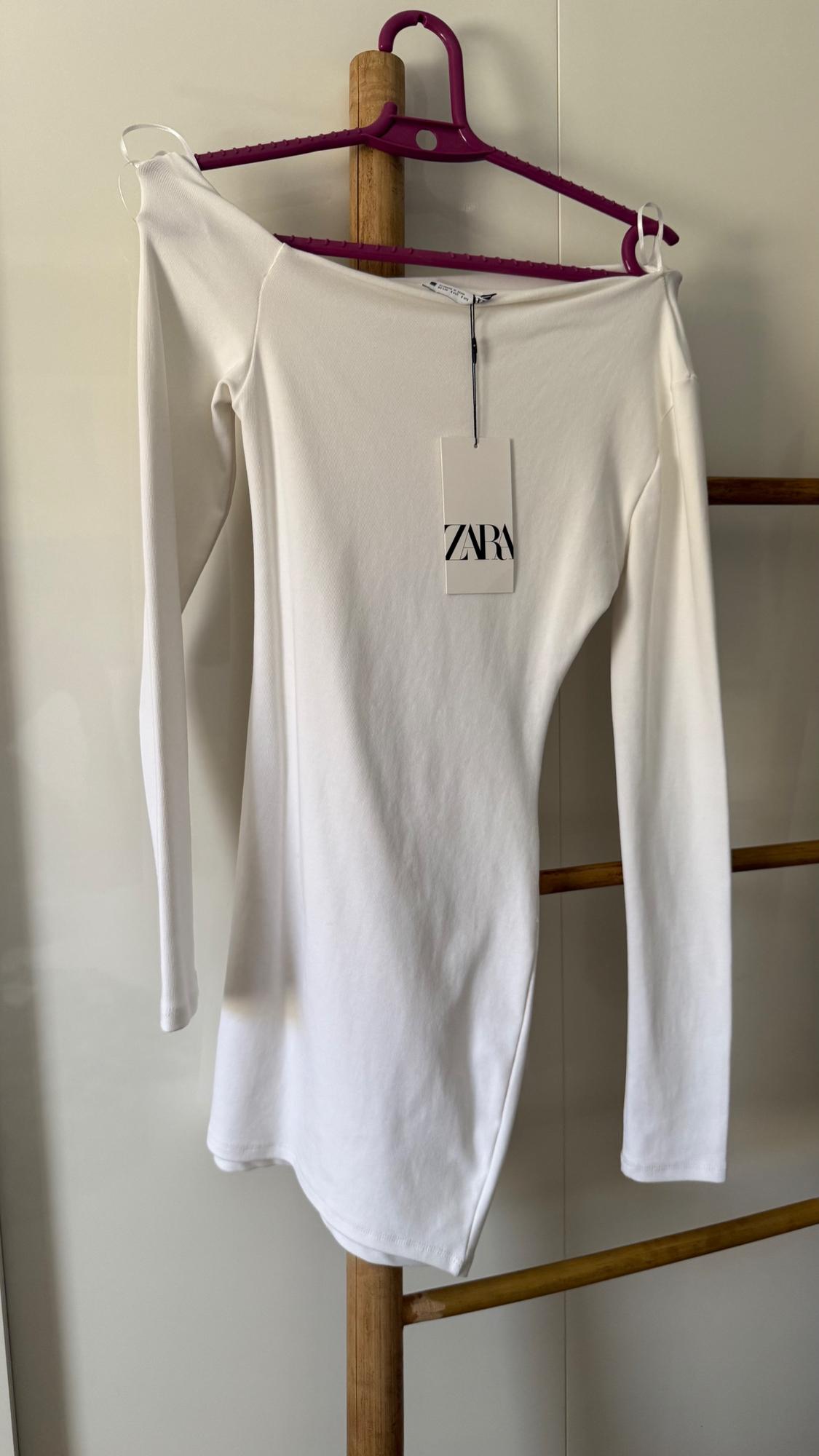 White long-sleeved dress hanging on a purple hanger. Visible label with the text "ZARA". White background.