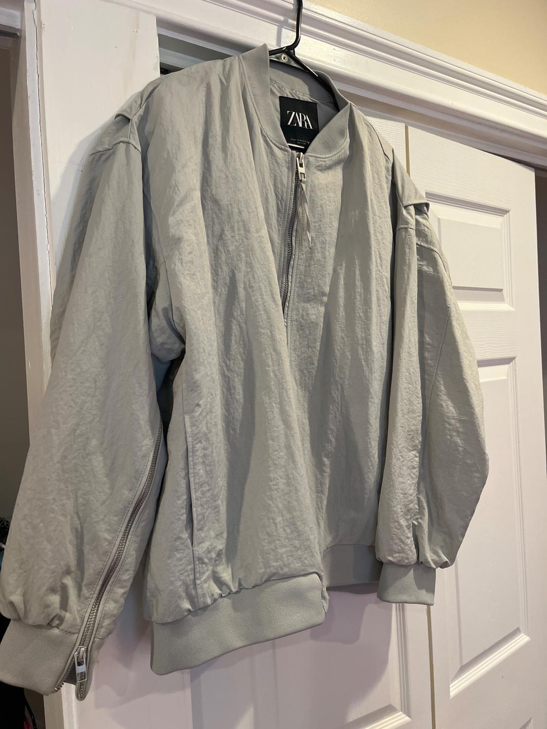Light grey jacket hanging on a white door, with a front zip and elasticated cuffs. Visible label with the text "ZARA".