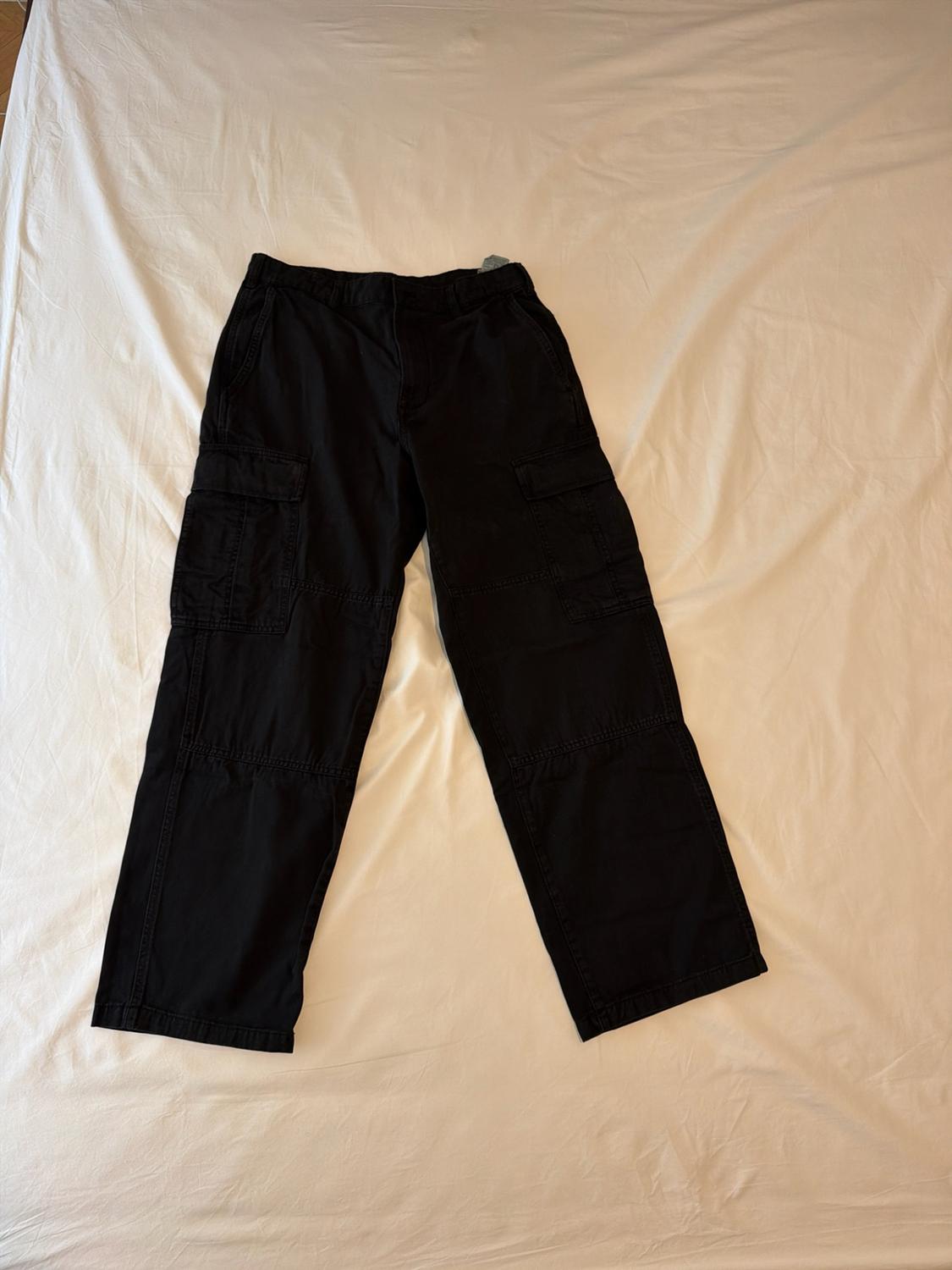 Black cargo trousers with side pockets, laid out on a crumpled white sheet in a casual-style composition.