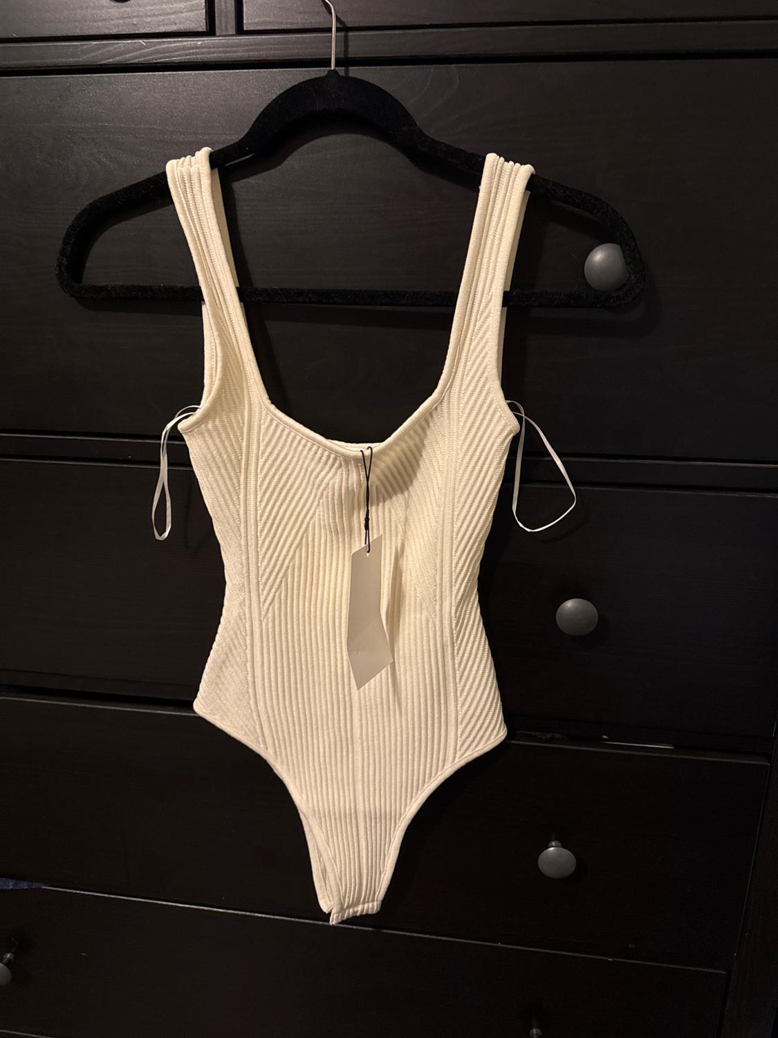 White ribbed bodysuit hanging on a black hanger in front of a dark piece of furniture. Tag hanging from the center of the bodysuit.