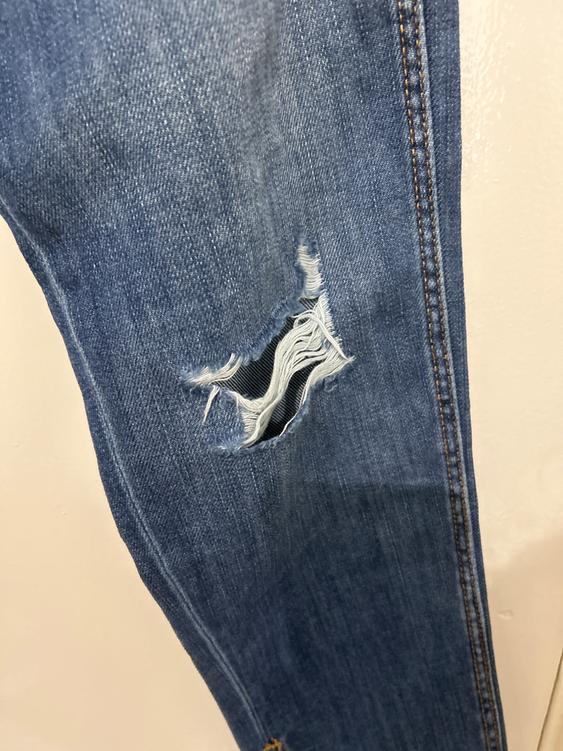 Blue denim jeans leg with a large rip at the top, showing frayed threads and a dark background.