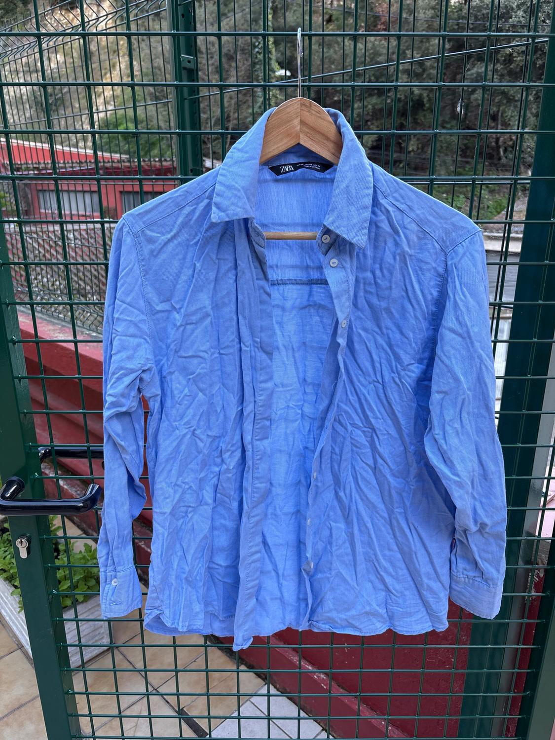Creased blue shirt hanging on a wooden hanger over a green railing, with a background of vegetation and a red structure.