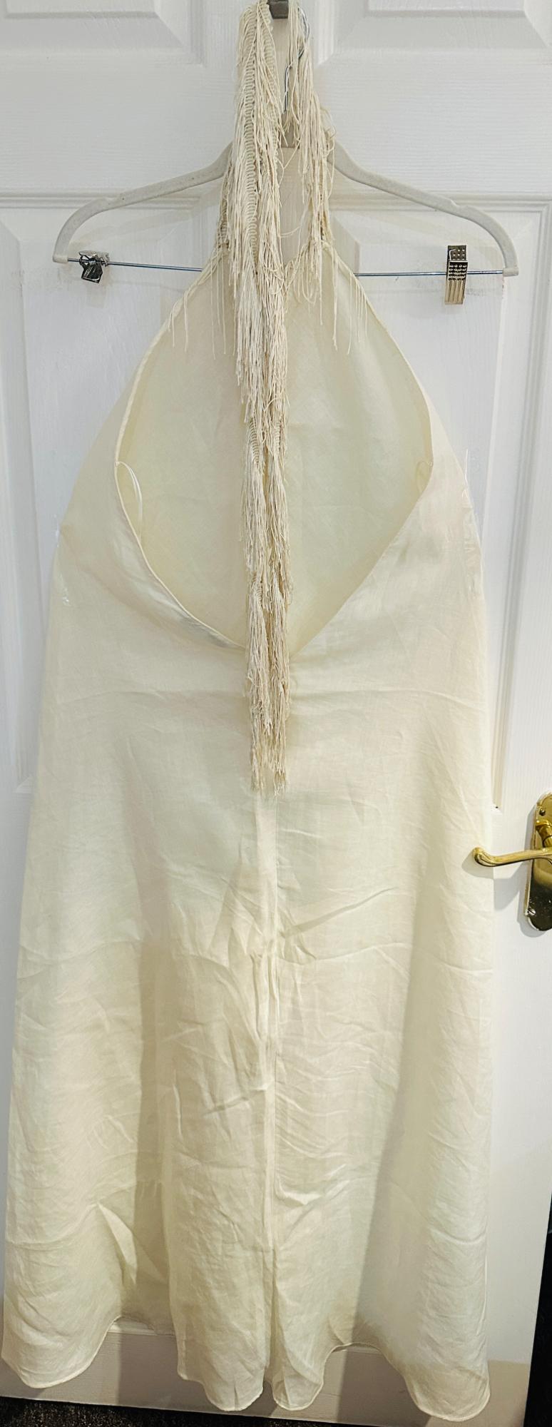 Cream halter-neck dress with an open back and long fringing, hanging on a hanger over a white door with a gold handle.