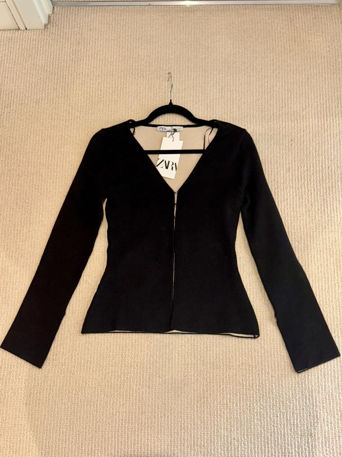 Black, long-sleeved V-neck jacket, hanging on a hanger over a beige rug. Visible label with text "ZARA".