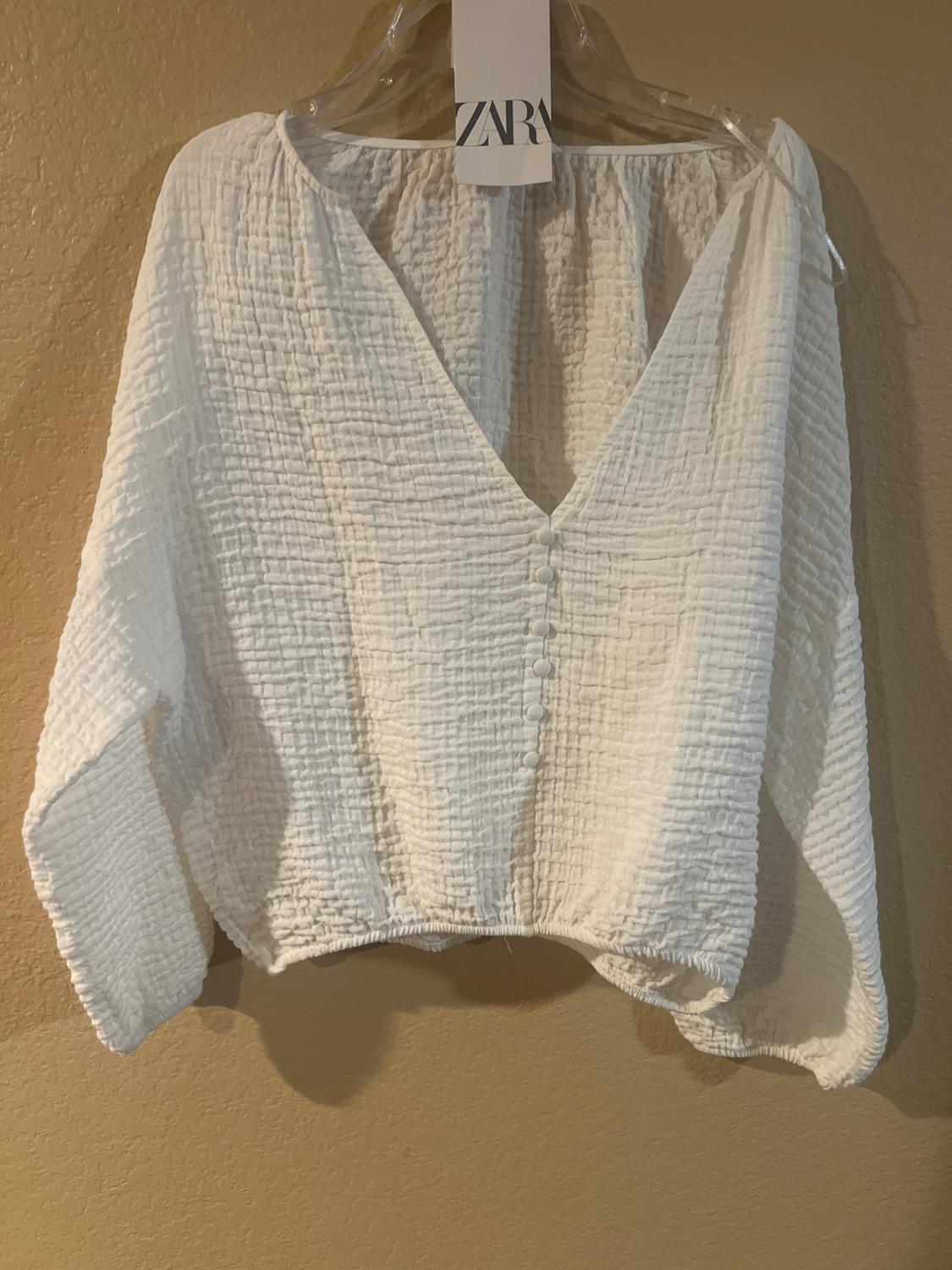 White crinkle textured blouse with a V-neck and front buttons, hanging on a hanger with a visible ZARA tag.