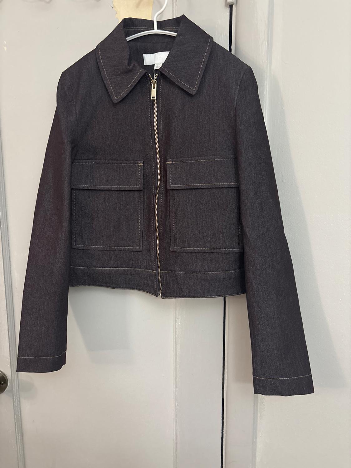 Dark grey jacket with a zip, shirt collar and two front flap pockets, hanging on a coat hanger on a white door.