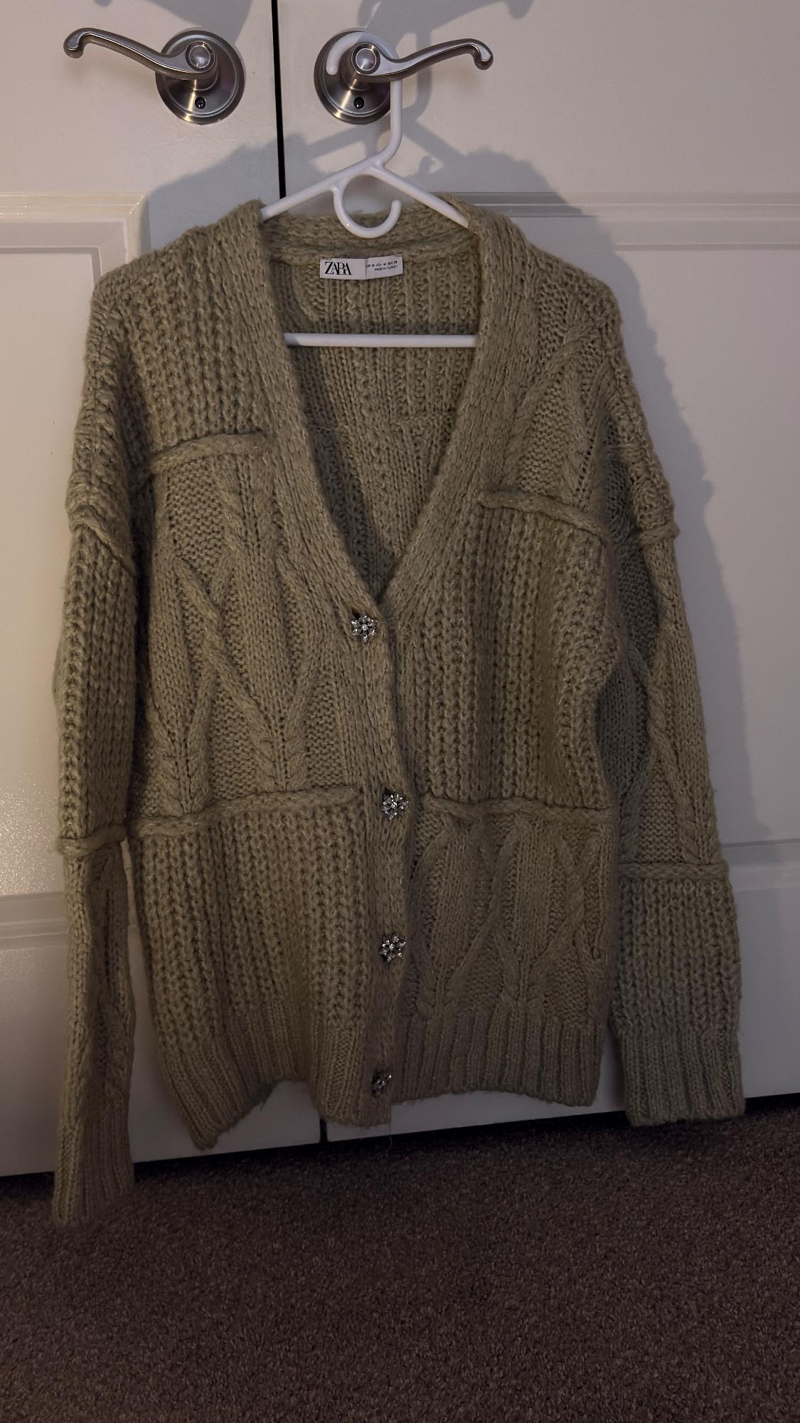 Chunky knit beige cardigan hanging on a white hanger. Detail of shiny buttons and visible Zara tag. White doors in the background.