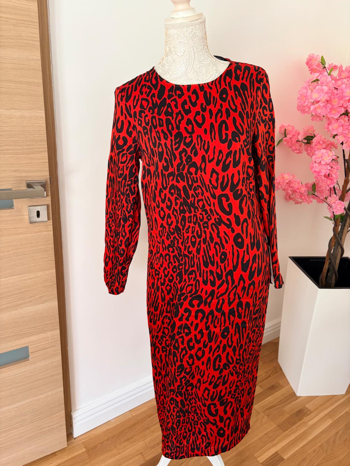 Μανεκέν με κόκκινο φόρεμα με animal print σε δωμάτιο με ξύλινο πάτωμα και ροζ λουλούδια σε λευκή γλάστρα.