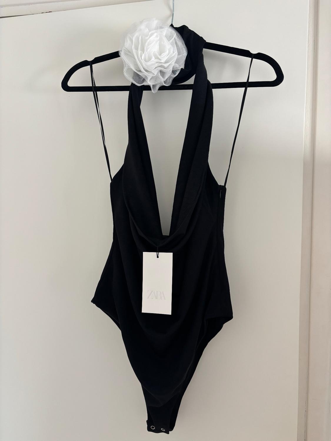 Black bodysuit with draped neckline on hanger, white floral embellishment on the shoulder. Label with text "ZARA". White background.
