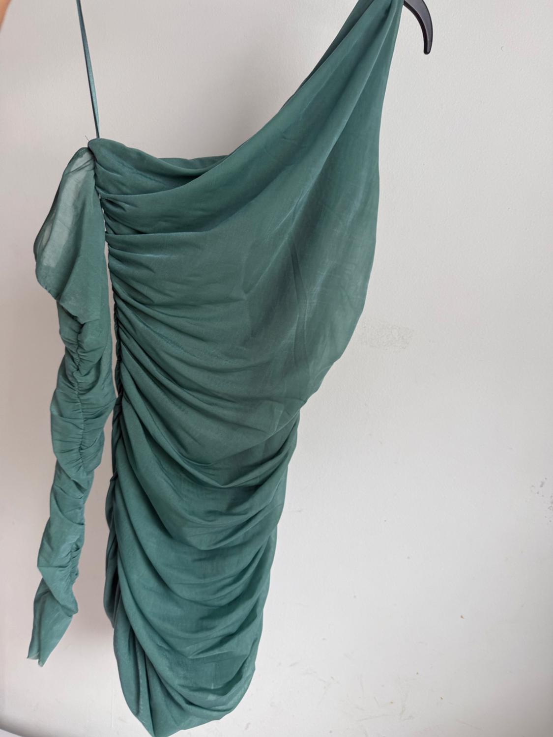 Asymmetrical green dress with draping and long gathered sleeves, hanging on a black hanger against a white wall.