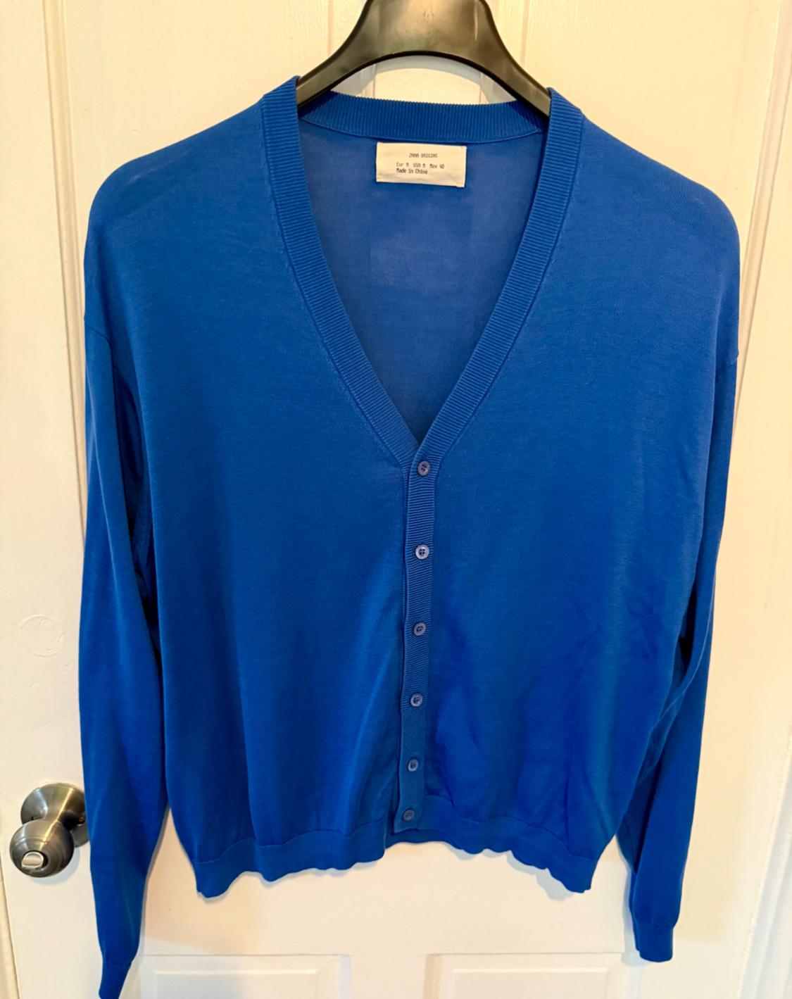 Blue cardigan with buttons, hanging on a black coat hanger in front of a white door. Label visible on the neck.