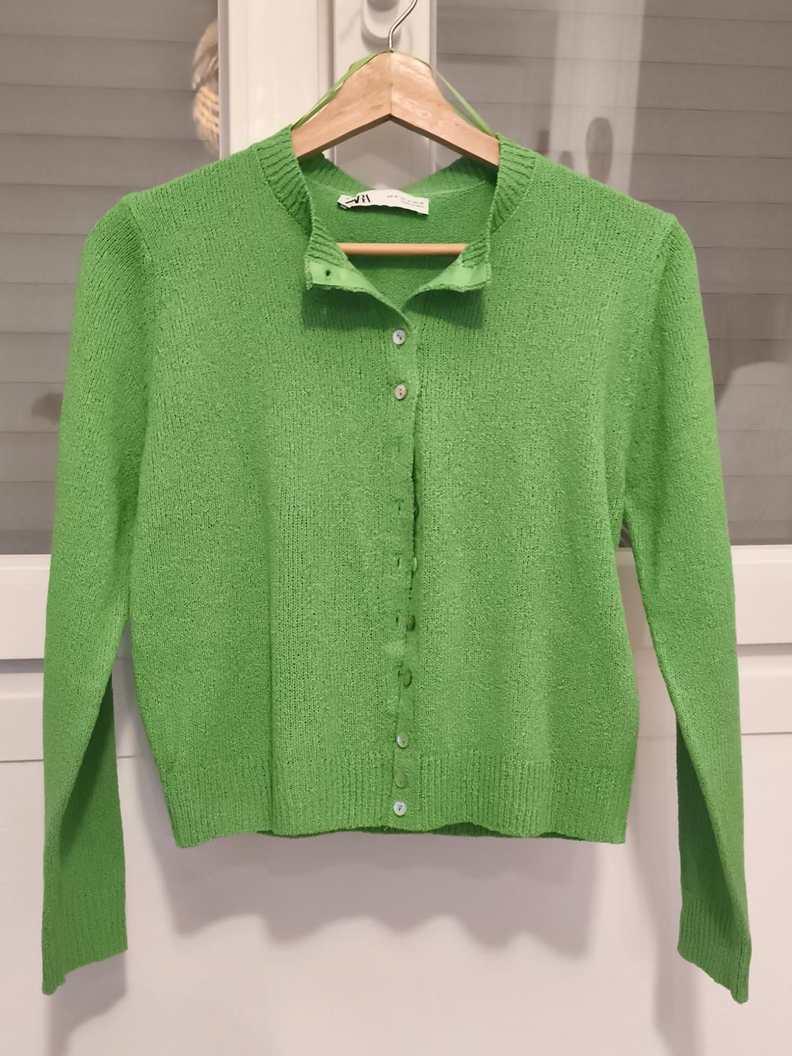 Green knitted cardigan hanging on a wooden hanger in front of a window with blinds.