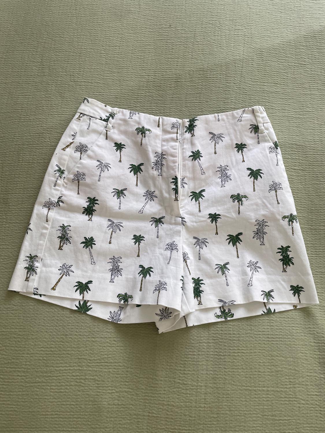 White shorts with a green palm tree print on a textured light green fabric background. Relaxed, summery style.