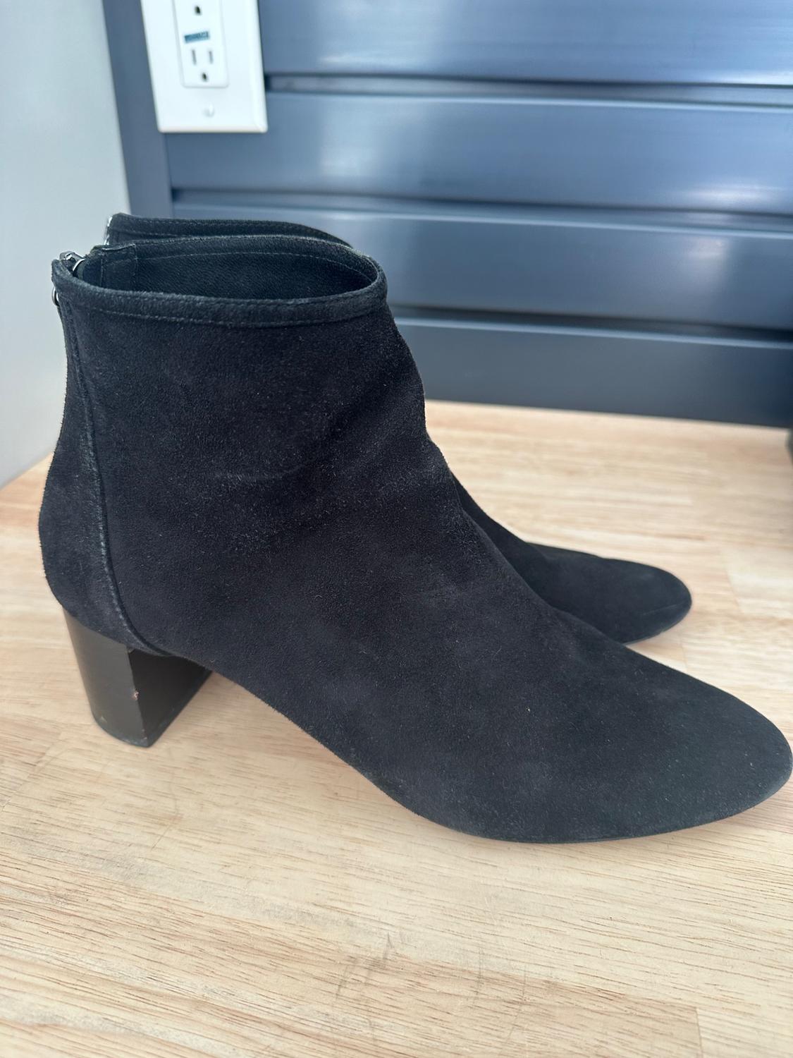 Black mid-heel ankle boots on a light wooden surface, with a gray paneled background and visible electrical outlet.