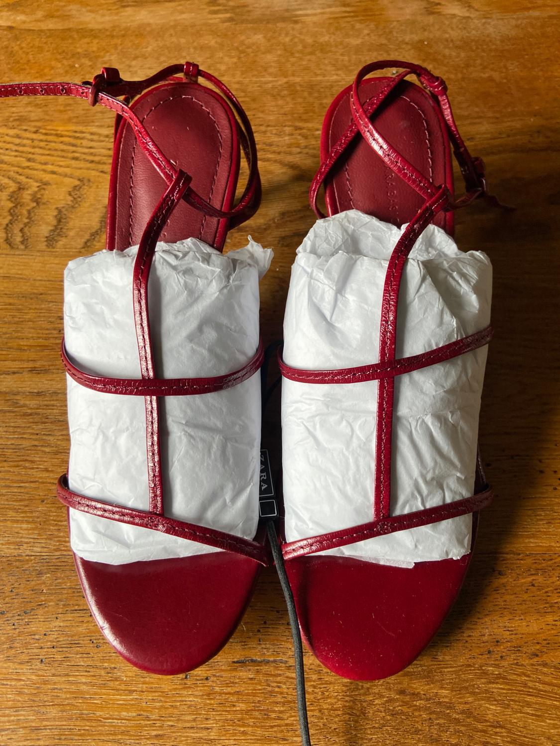 Pair of red strappy high-heeled sandals, stuffed with white paper,
on a wooden surface. Label with the text ZARA.