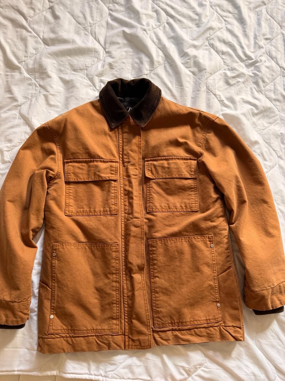 Brown jacket with a dark corduroy collar and four front pockets, photographed on a white quilted duvet.