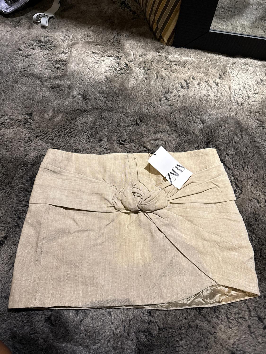 Beige miniskirt with a front knot on a textured gray rug. Tag with the text ZARA.