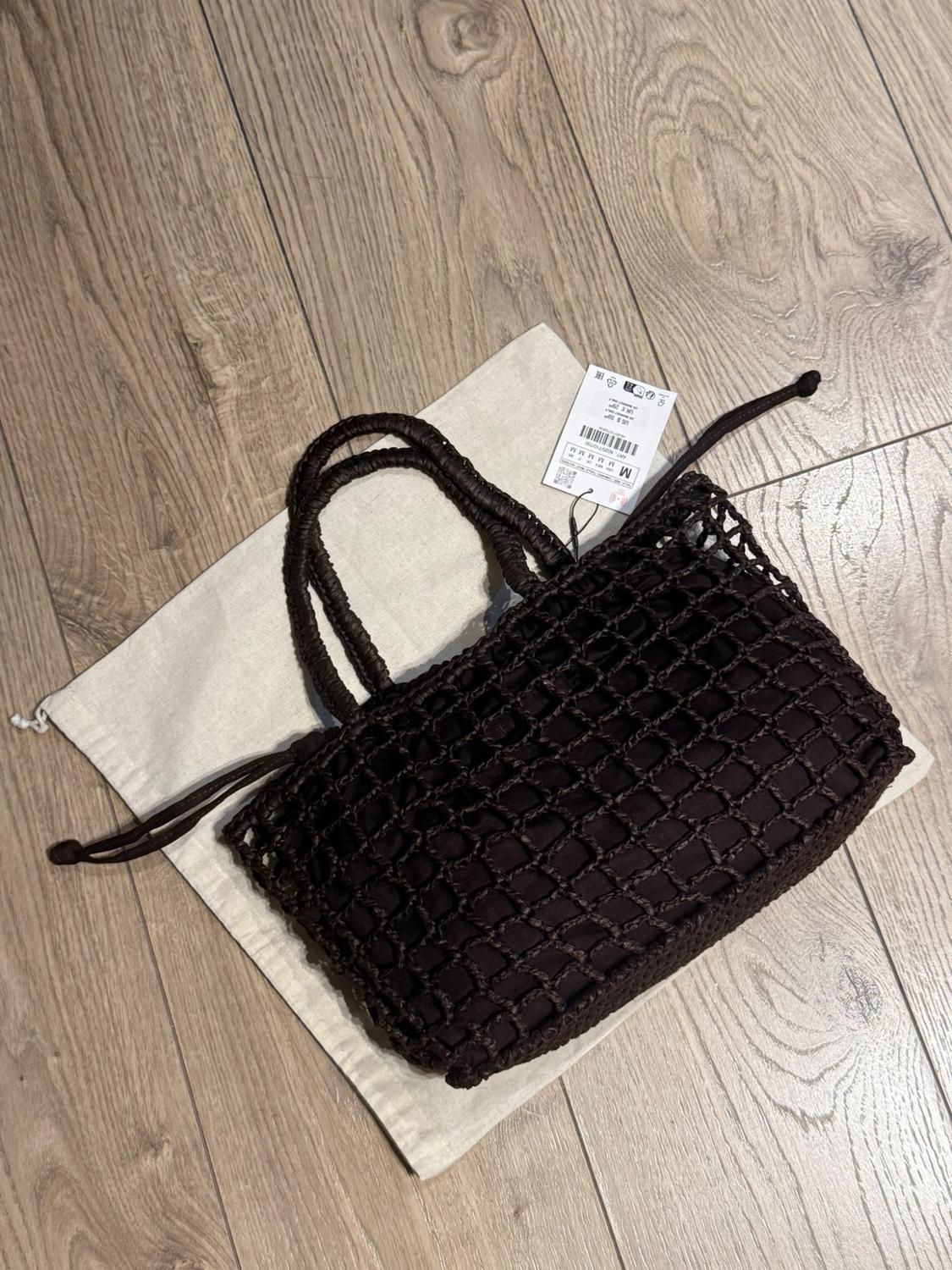 Dark brown mesh bag with handles and drawstrings, resting on a fabric bag on a wooden floor. Label with the text M M M M M M.