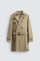 Front view of beige double-breasted trench coat with adjustable belt and side pockets.