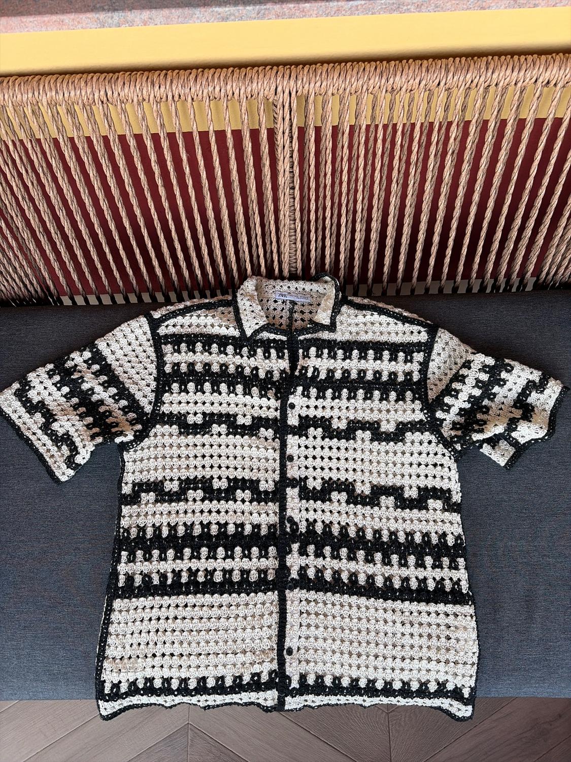 Black and white crochet shirt with a geometric pattern, over a background of braided rope and a grey sofa.
