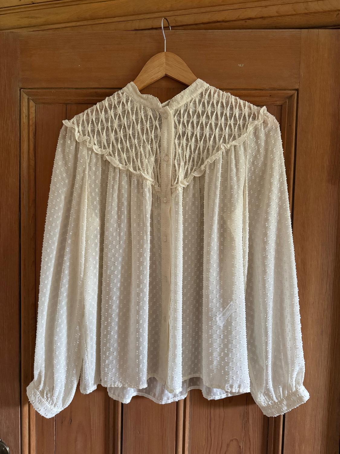 White lace blouse with a high neck and long sleeves, hanging on a wooden hanger over a wooden door.