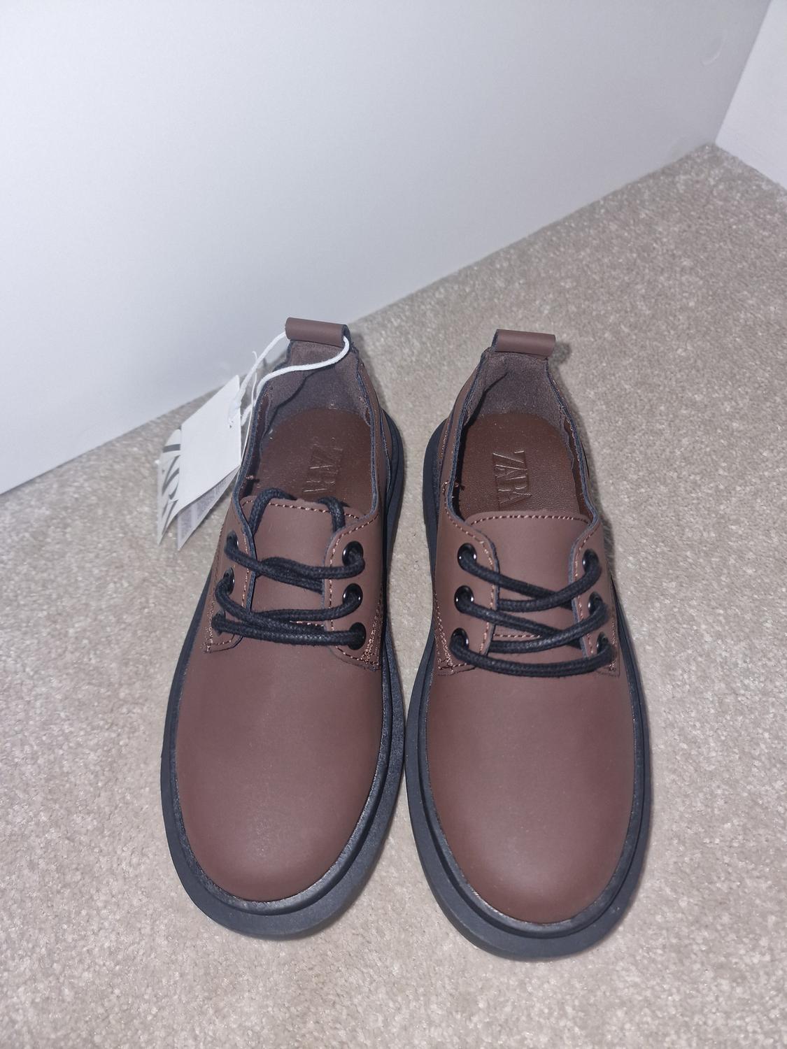 Brown shoes with black laces on a beige carpet. White labels visible. White background.
