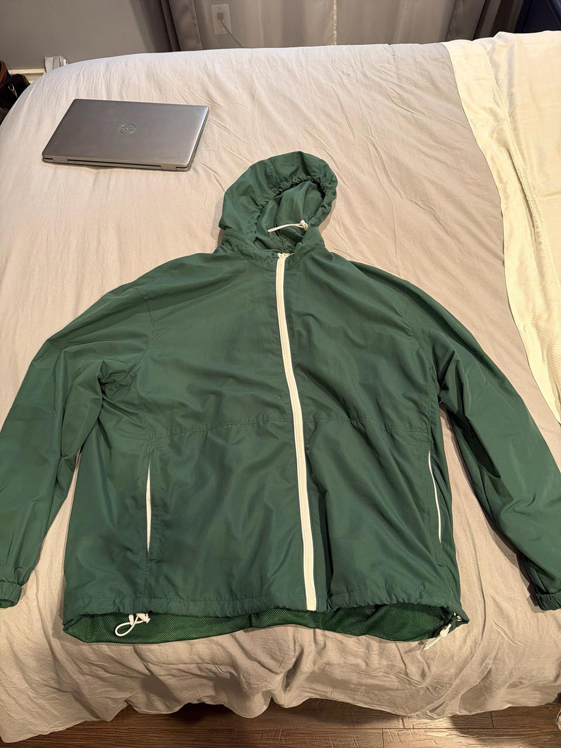Green hooded jacket with a white zip on a bed with light-coloured sheets. A closed laptop in the background adds a relaxed feel.