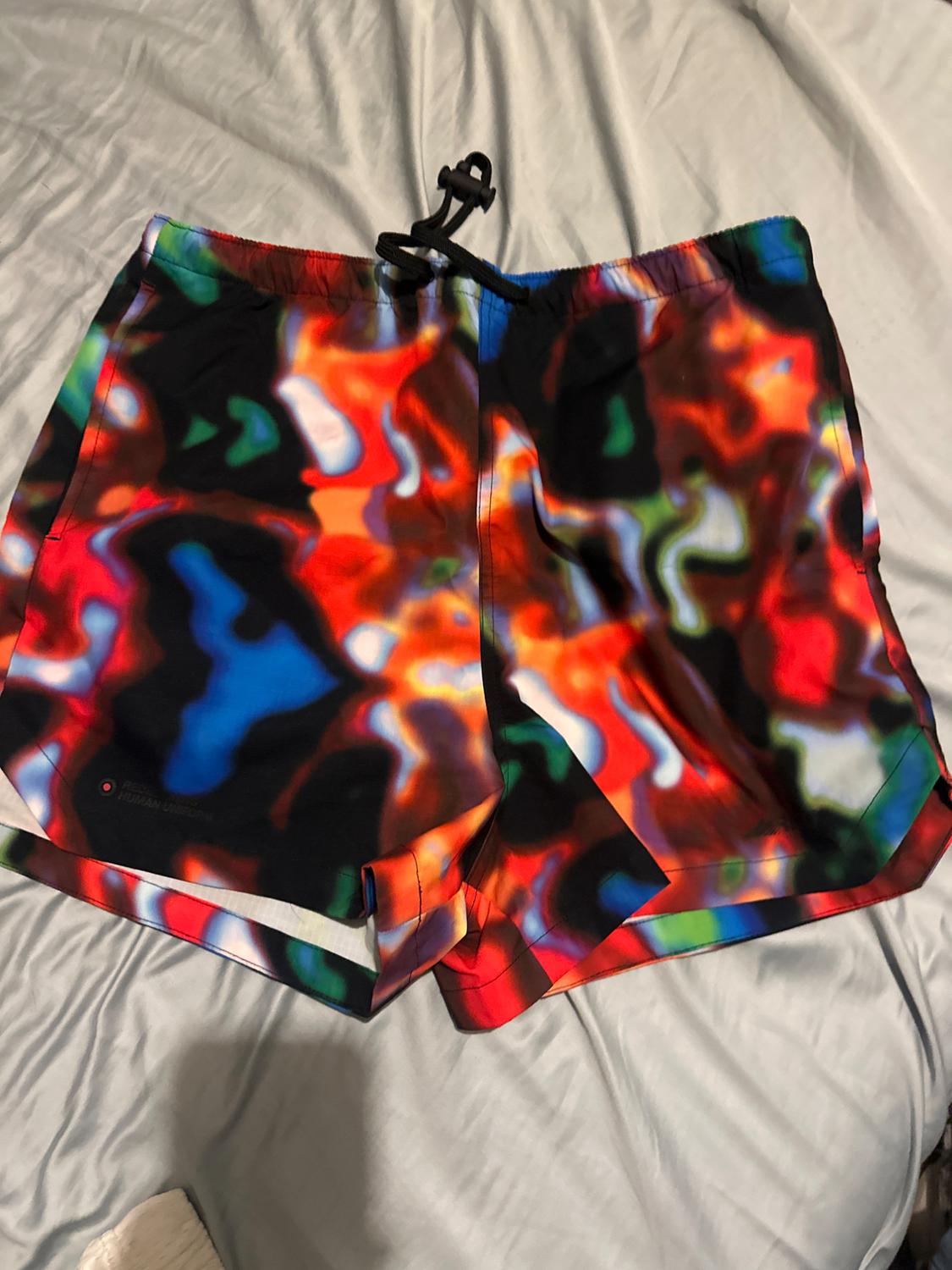 Shorts with a vibrant, multicoloured abstract print and a black drawstring, laid on a crumpled sheet. Text: REBEL HUMAN UNIFORM.