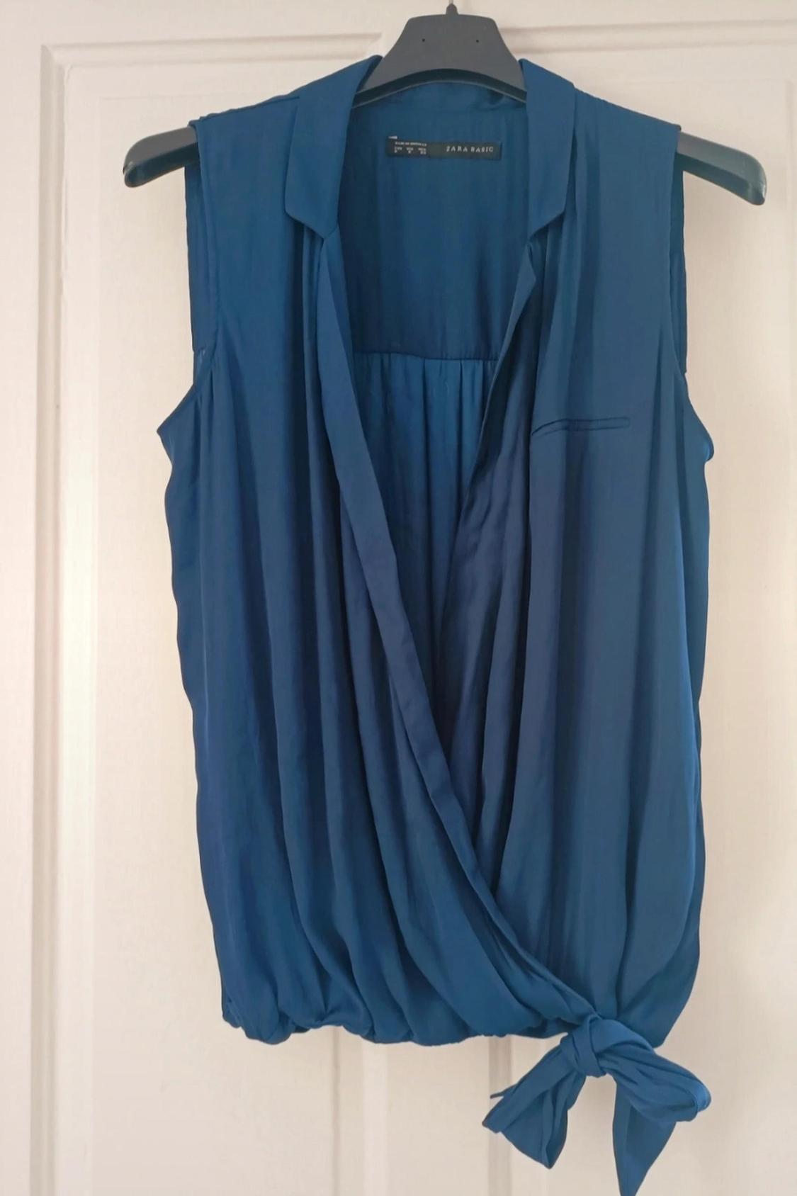 Front view of a blue sleeveless blouse with a classic collar and a knot at the hem.