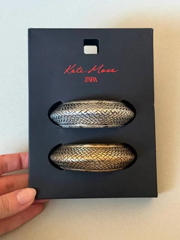 A black box with two textured gold bracelets. The box reads: "Kate Moss ZARA". A hand holds the box.