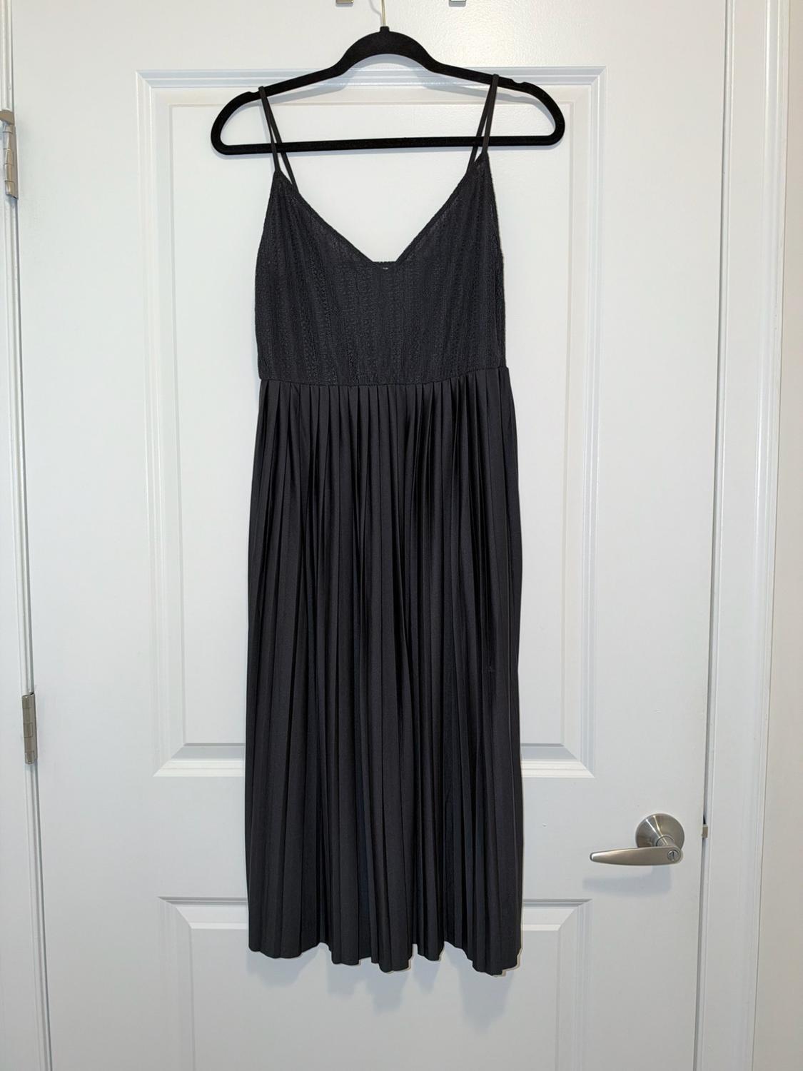 Black spaghetti-strap dress with a pointed neckline, textured bodice, and a pleated skirt, hanging on a hanger over a white door.