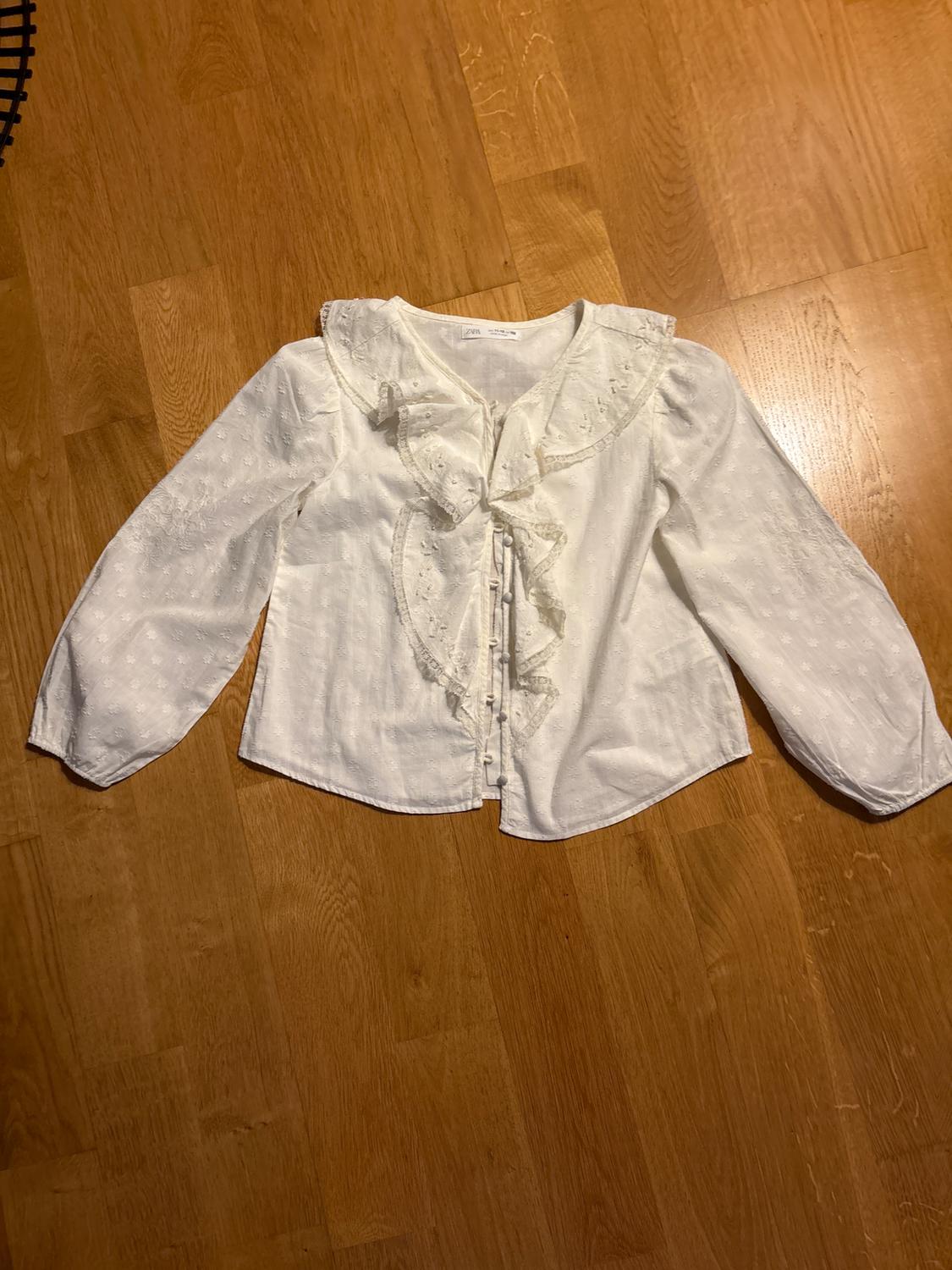 White long-sleeved blouse with ruffles and embroidered details, on a light wooden floor.