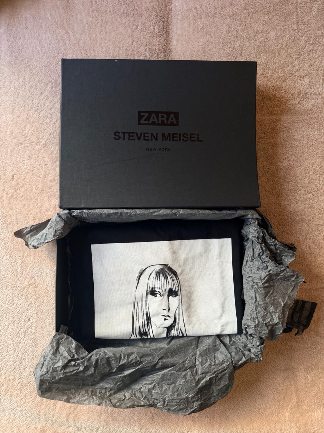 Open black box with ZARA STEVEN MEISEL NEW YORK text. Contains paper and a black garment with an illustrated portrait on a white background.