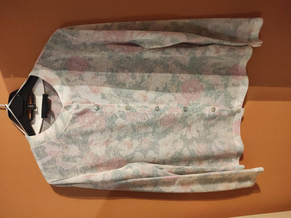 Knitted cardigan with floral print in pastel shades, hanging on a black hanger against an orange wall.
