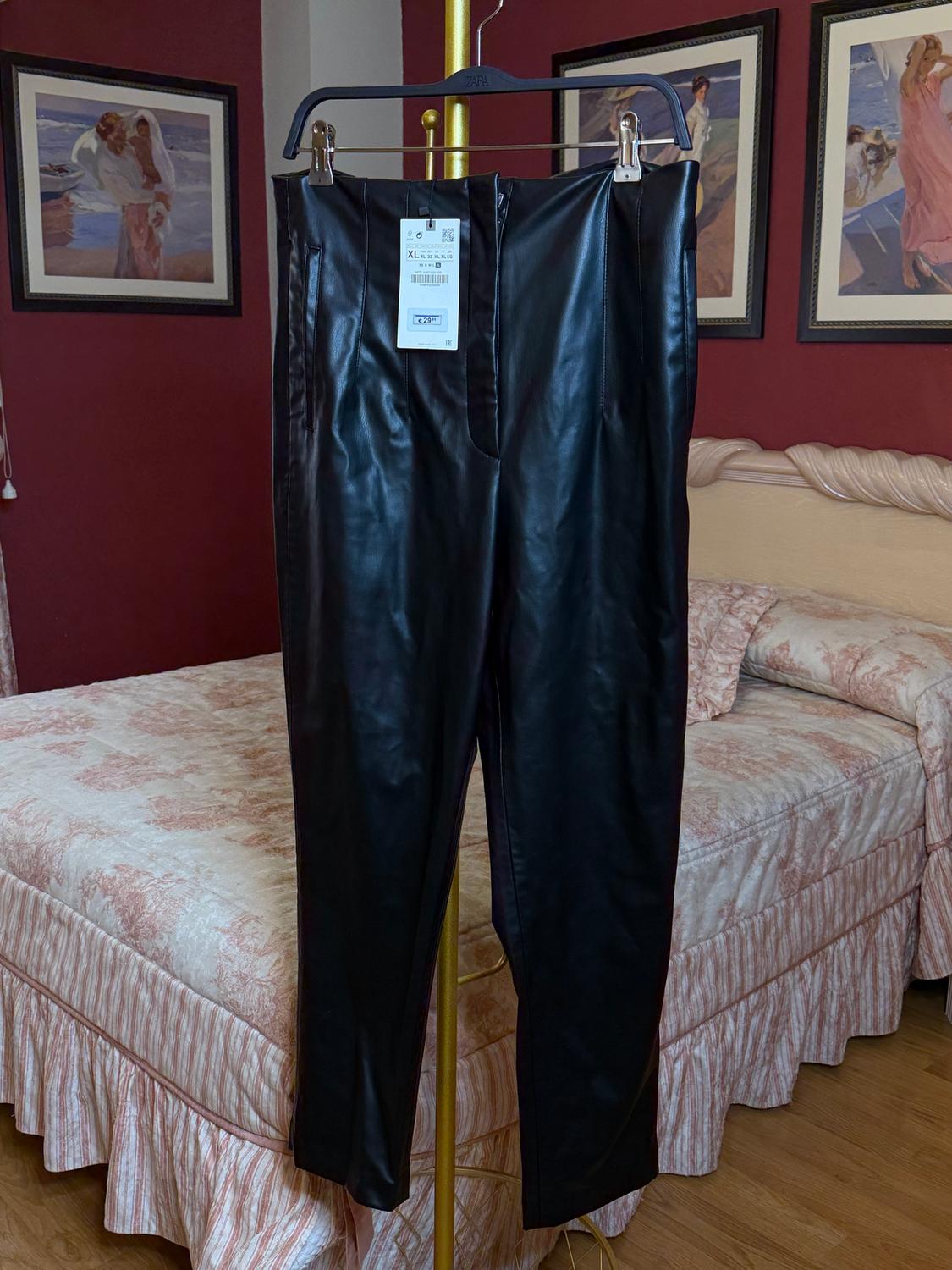 Black trousers hanging on a gold hanger in a bedroom with red walls and pictures. Visible tag with size XL.